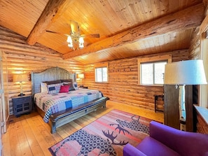 A log cabin bedroom with a bed, a ceiling fan, a window, and a lamp.