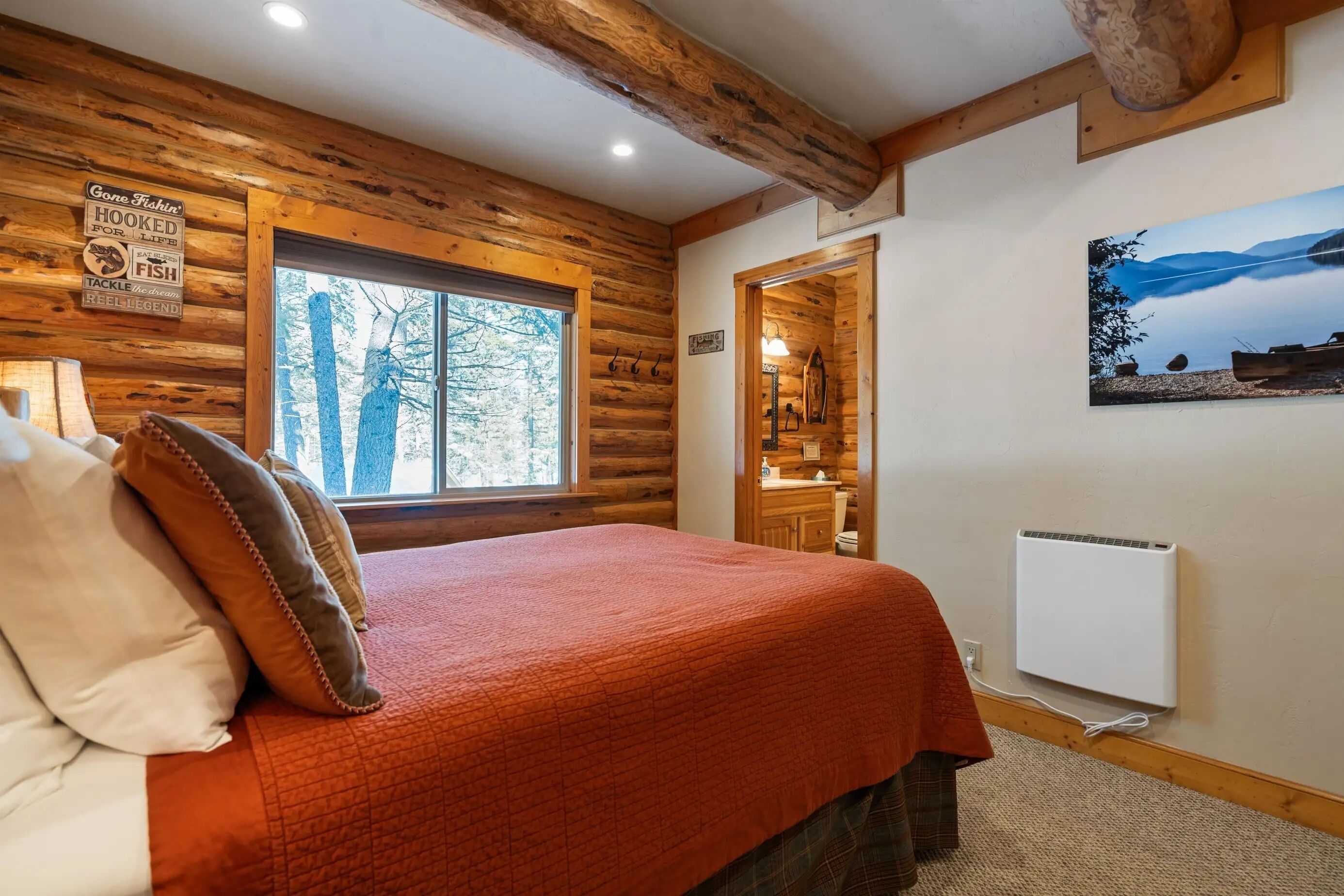 A log cabin bedroom with a large window, a bed with a red blanket, a wooden ceiling, and a painting on the wall.