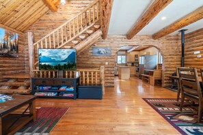 A log cabin interior with a wooden staircase, a television, a fireplace, and a kitchen area.