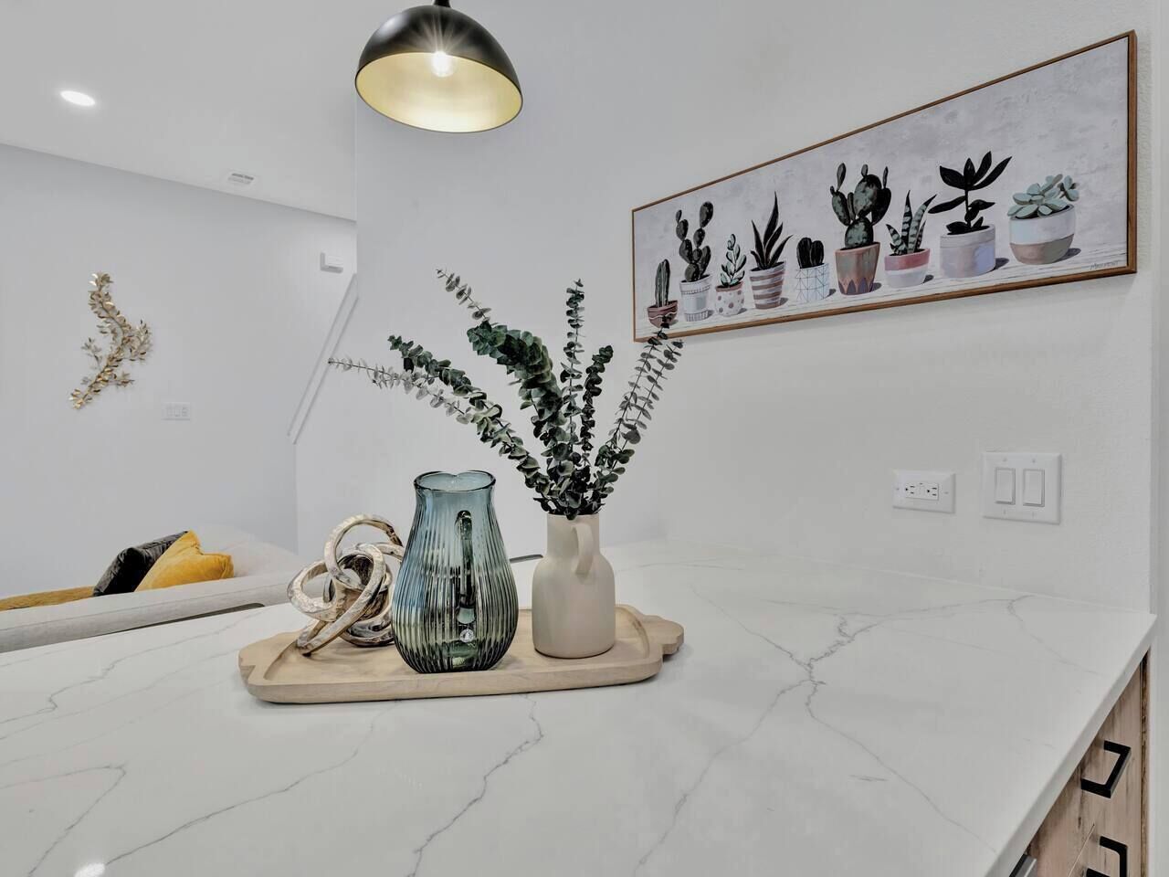 A modern kitchen with a marble countertop, a pendant light, a vase with plants, and a framed plant illustration on the wall.