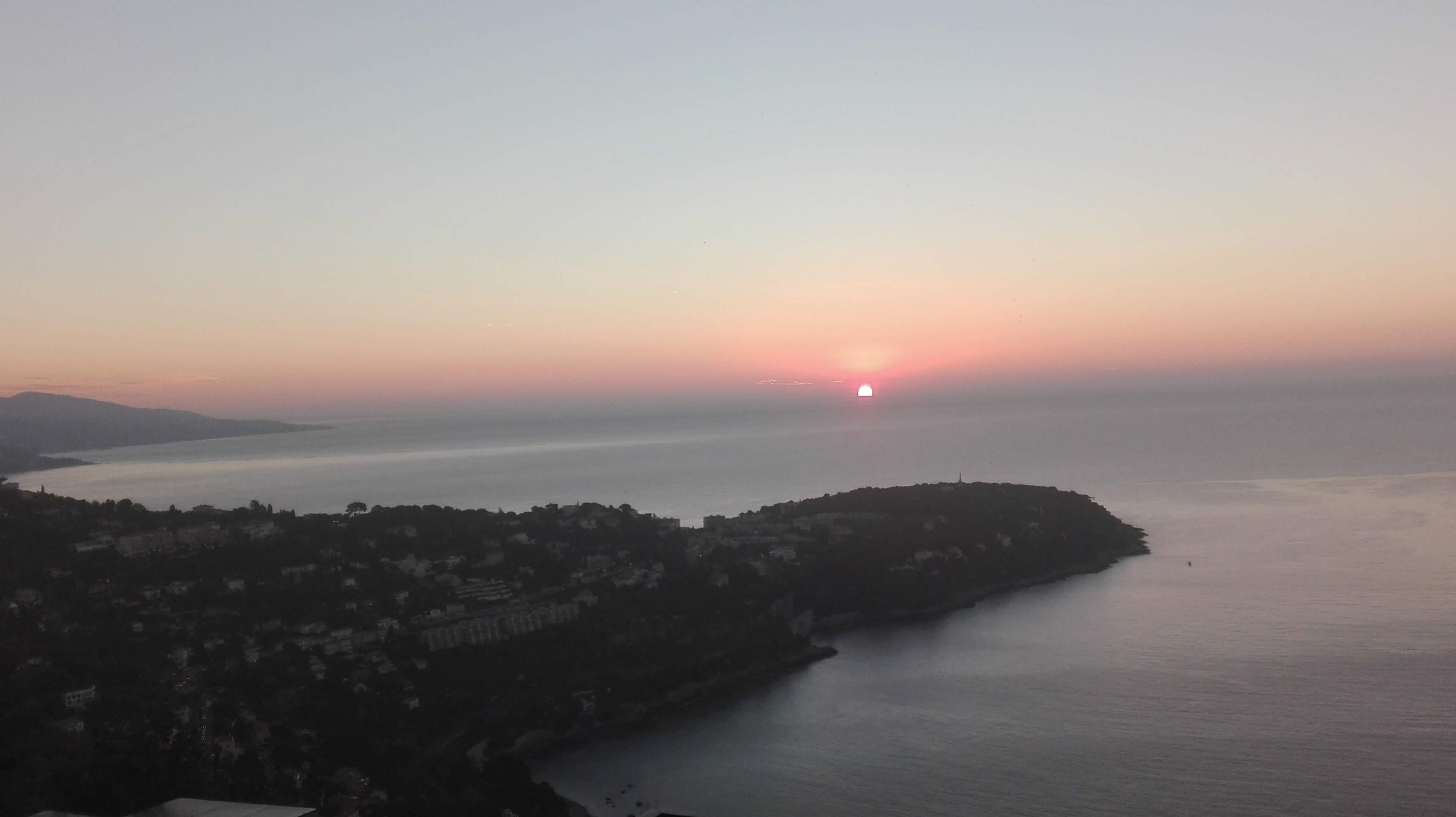 A coastal landscape with a sunset over the sea, a hilly area with buildings, and a clear sky.