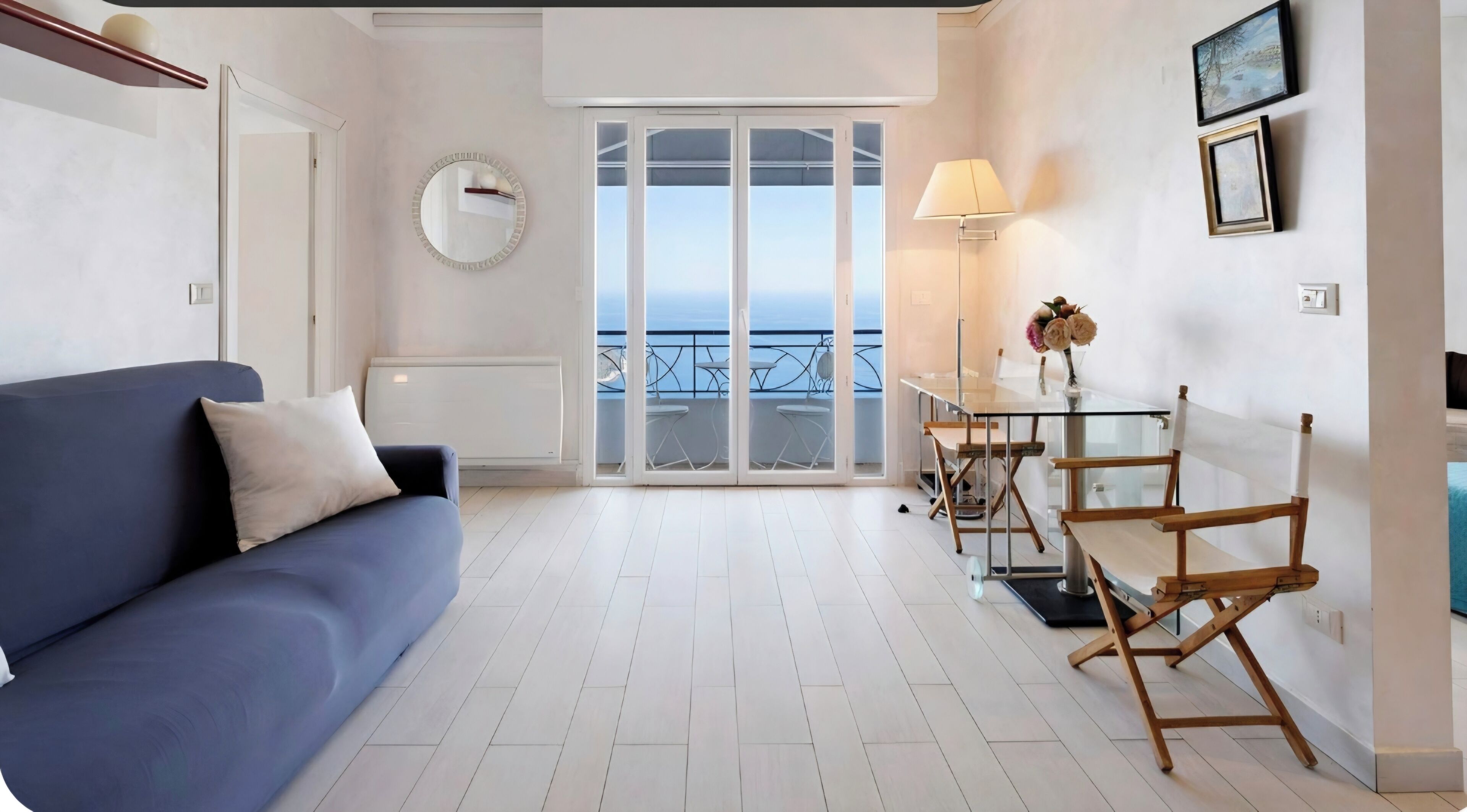Un salon moderne avec un canapé bleu, un miroir rond, une table à manger avec des chaises et une vue sur la mer, visible à travers une porte vitrée.