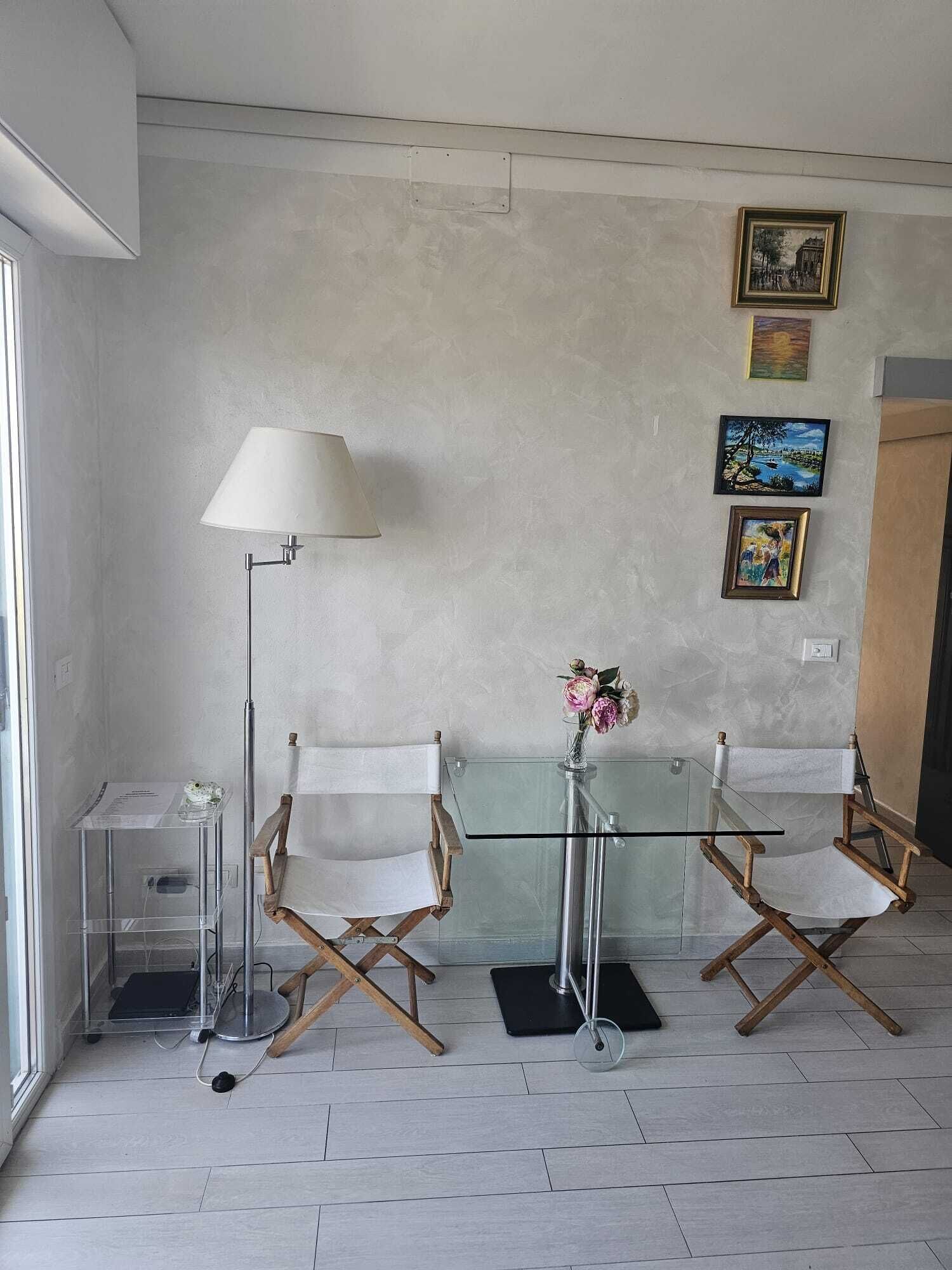 Una habitación con una mesa de vidrio, dos sillas de madera, una lámpara de pie, una mesita con un florero y tres cuadros en la pared.