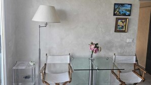Una habitació amb taula de vidre, dues cadires de fusta, una làmpada de peu, una taula petita amb un ram de flors i tres quadres al mur.