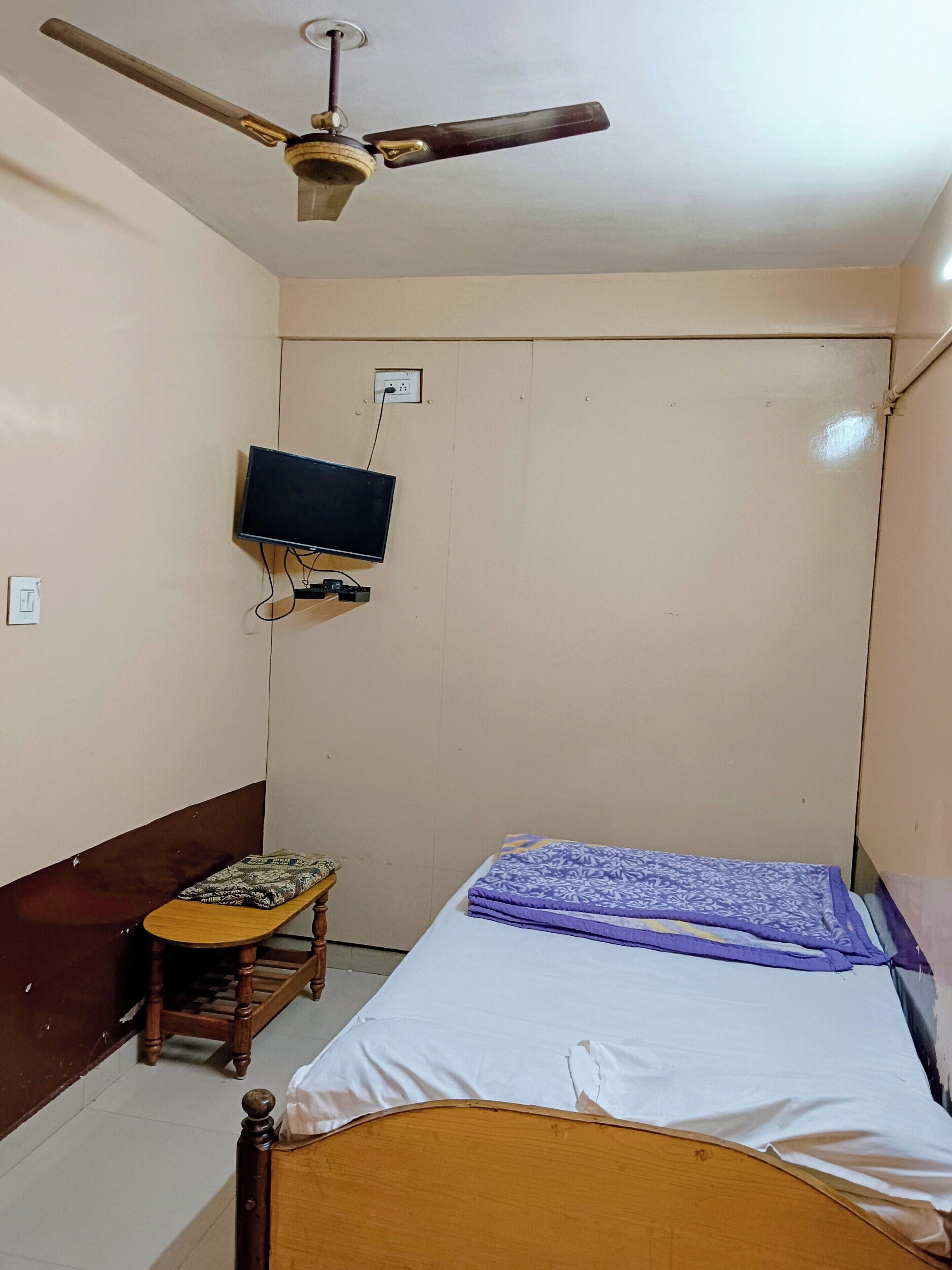 Ein Schlafzimmer mit einem Bett, einem Deckenventilator, einem an der Wand montierten Fernseher und einem kleinen hölzernen Hocker.