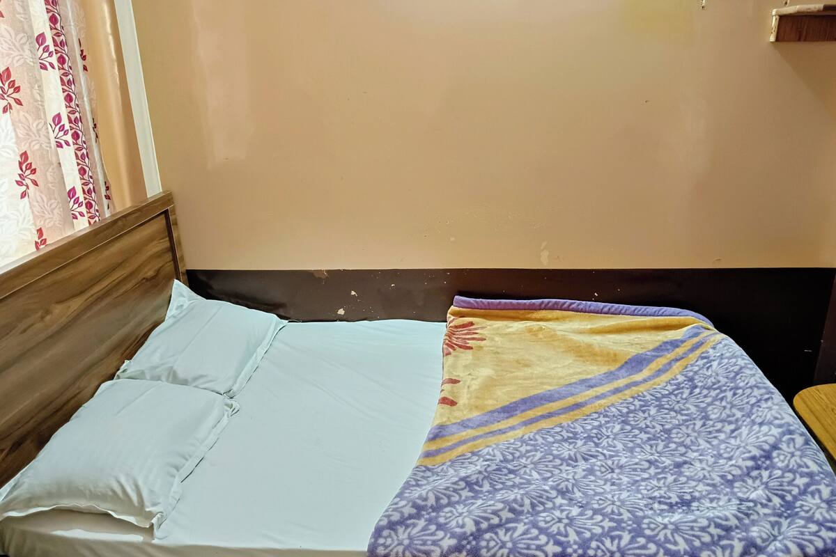 A bed with a white sheet and a purple patterned blanket, a wooden headboard, and a window with floral curtains.