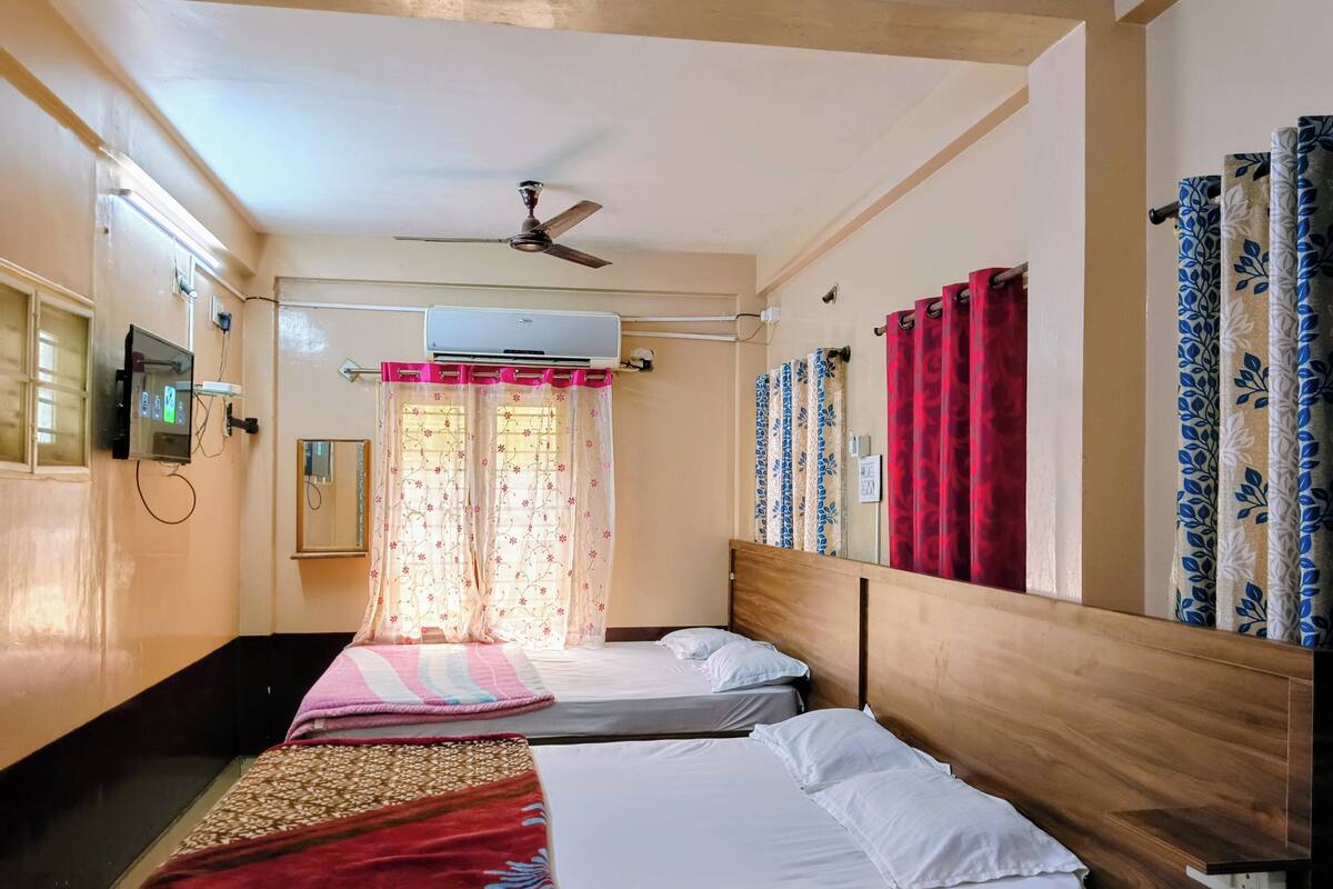 A hotel room with two single beds, a ceiling fan, an air conditioning unit, a TV mounted on the wall, and a window with floral curtains.