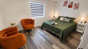Een slaapkamer met een bed, twee stoelen, een klein tafeltje, een raam met jaloezieën en wanddecoratie.