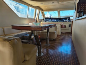 A boat interior with a dining table, cushioned seating, a control panel, and a view through the windows.