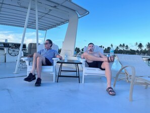 Two people relaxing on a boat deck with lounge chairs, a steering wheel, and a table with a water bottle.