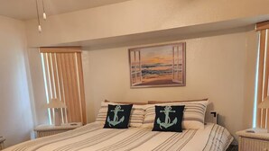Un letto con lenzuola a righe, due cuscini con motivi a ancora, un comodino in vimini, un pouf intrecciato, un quadro raffigurante un tramonto sull'oceano e una finestra con persiane verticali.