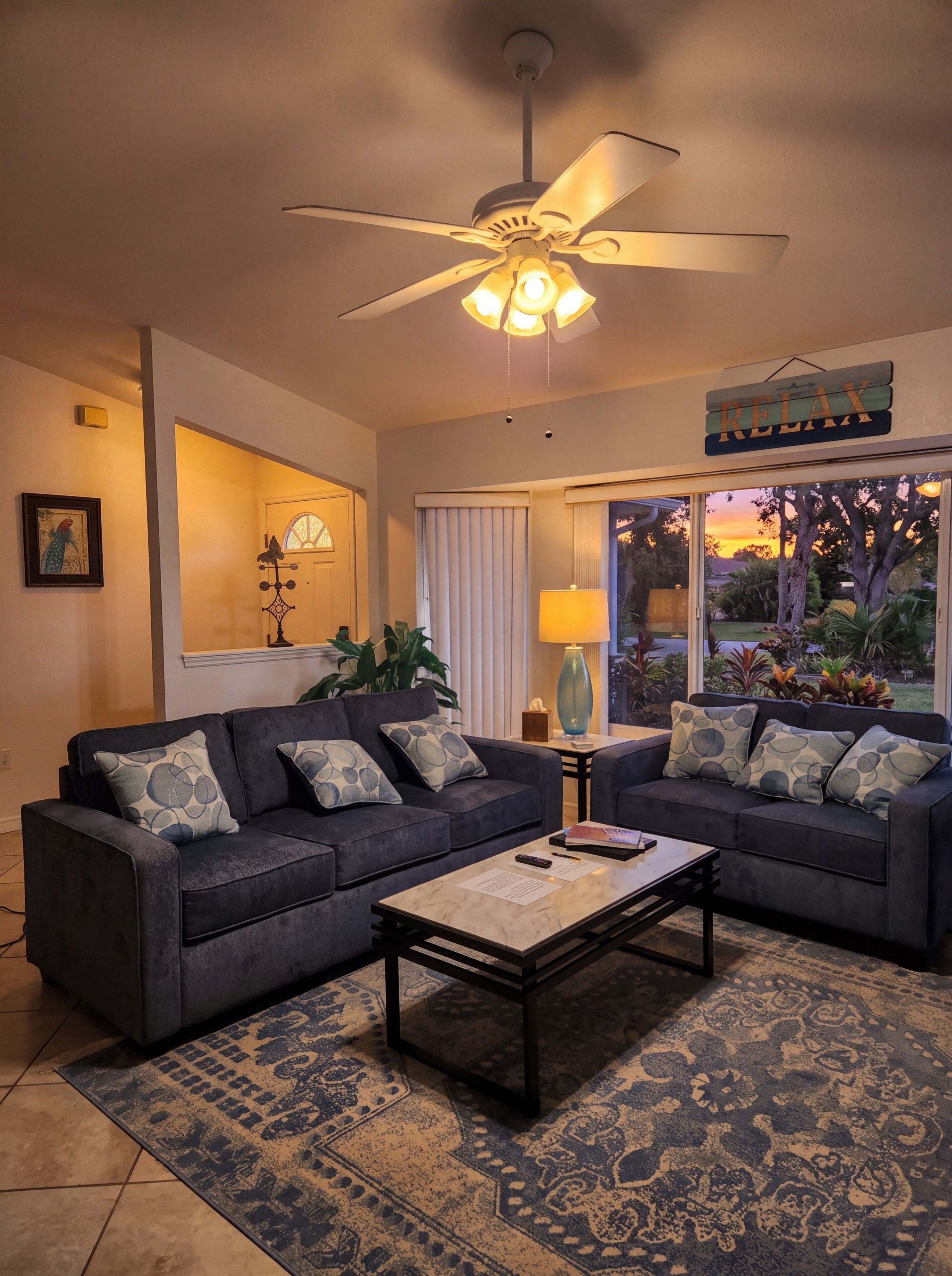 A living room with two sofas, a coffee table, a ceiling fan, a window with curtains, and a view of the outdoors.