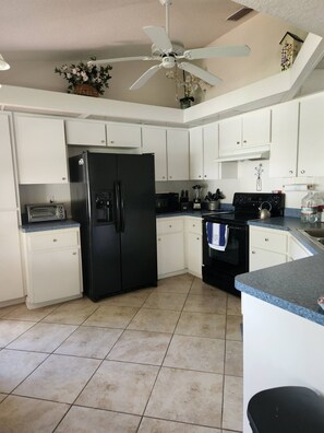 A kitchen with white cabinets, a black refrigerator, a microwave, a stove, and a ceiling fan. - Miss Ellie's Retreat (Port Charlotte)