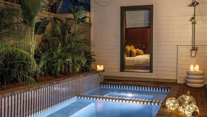 Een zwembad met een houten terras, omringd door weelderige begroeiing en een gezellige kamer met een bed en nachtkastje.