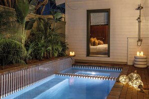 Ein Schwimmbecken mit Holzterrasse, umgeben von üppiger Vegetation und ein gemütliches Zimmer mit Bett und Nachttisch.