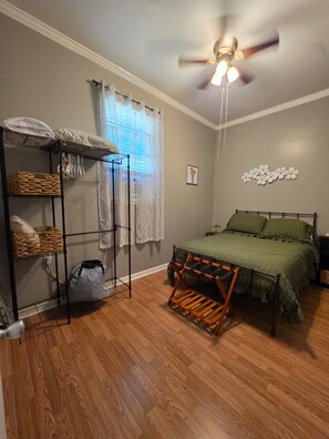 A bedroom with a bed, a ceiling fan, a window with curtains, a wall-mounted shelf with baskets, and a radiator. - Cozy home away from home (Matthews)