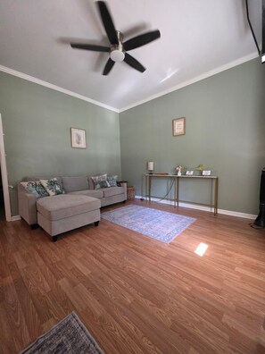 A living room with a ceiling fan, a grey sofa with decorative pillows, a small table with a lamp, and a blue rug. - Cozy home away from home (Matthews)