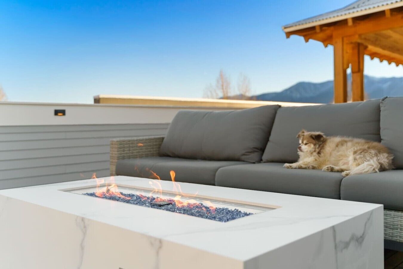 Ein Feuerkorb mit Flammen, ein graues Sofa und eine Holz-Pergola.