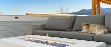 Ein Feuerkorb mit Flammen, ein graues Sofa und eine Holz-Pergola.