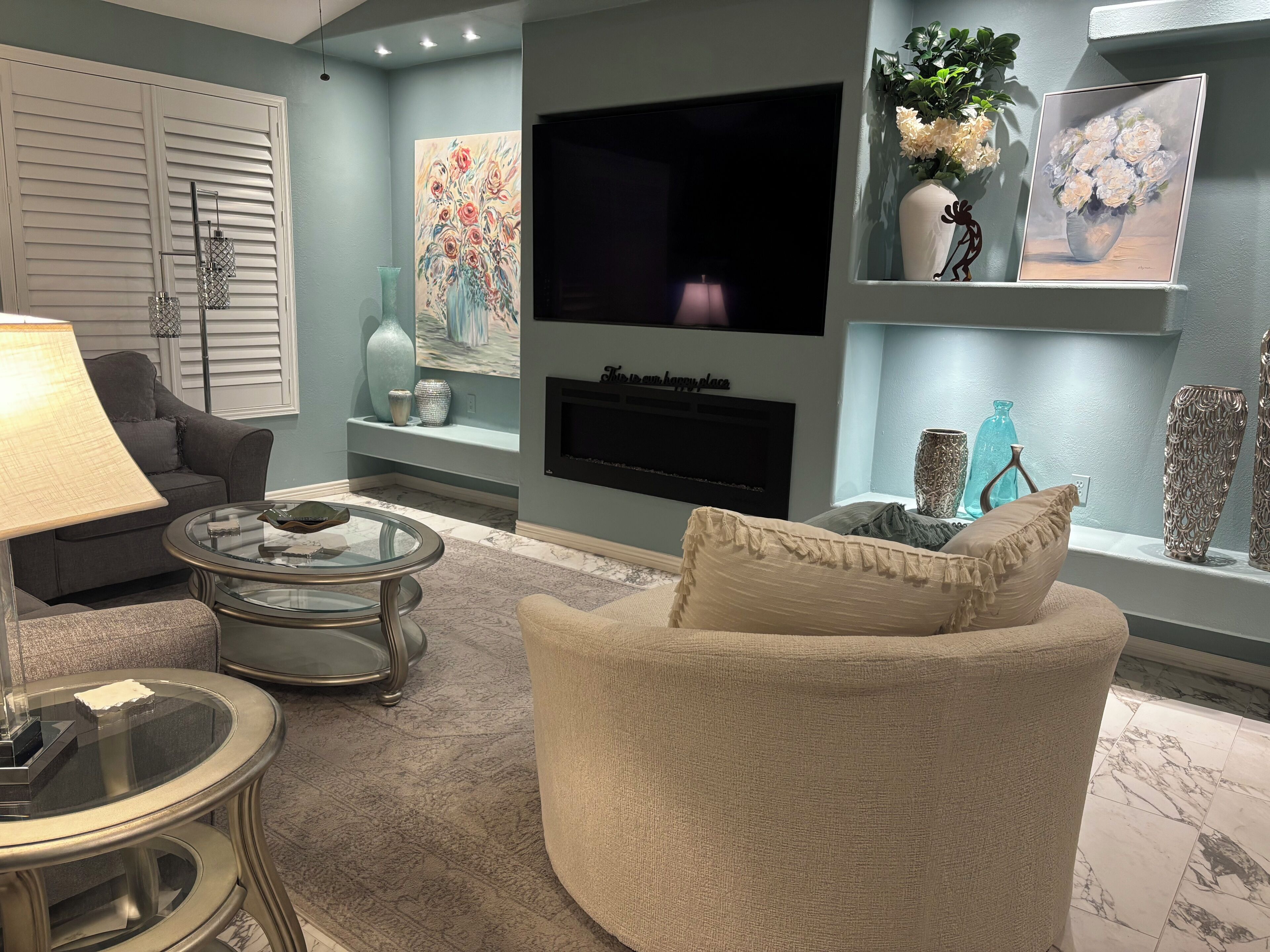 Un salon comprenant un canapé, une table basse en verre, un téléviseur fixé au mur et des objets décoratifs disposés sur des étagères.