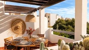 A round wooden table set for dining with a woven basket and flowers, surrounded by potted cacti, under a wooden pergola with a view of buildings and greenery. - Casa de la Alegria, 3BD house near Cabopino Beach (Marbella)