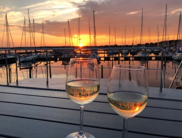 Dos copas de vino blanco sobre una mesa de madera, con un puerto deportivo y barcos al fondo.