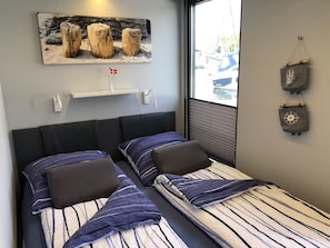 Une chambre avec deux lits, un tableau représentant des arbres, une fenêtre donnant sur des voitures garées et une étagère fixée au mur.
