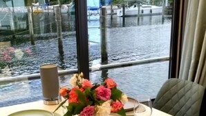 A dining table set with plates, cutlery, and a vase of flowers, with a view of boats through large windows.