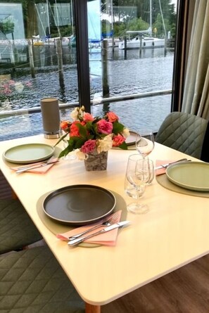 A dining table set with plates, cutlery, and a vase of flowers, with a view of boats through large windows.