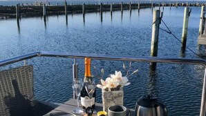 A table set for a meal with a bottle of champagne, a glass, a flower vase, a teapot, and a mug on a deck overlooking water.
