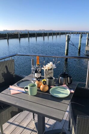 Una mesa preparada para una comida, con una botella de champán, una copa, un florero, una tetera y una taza, en una terraza con vistas al agua.