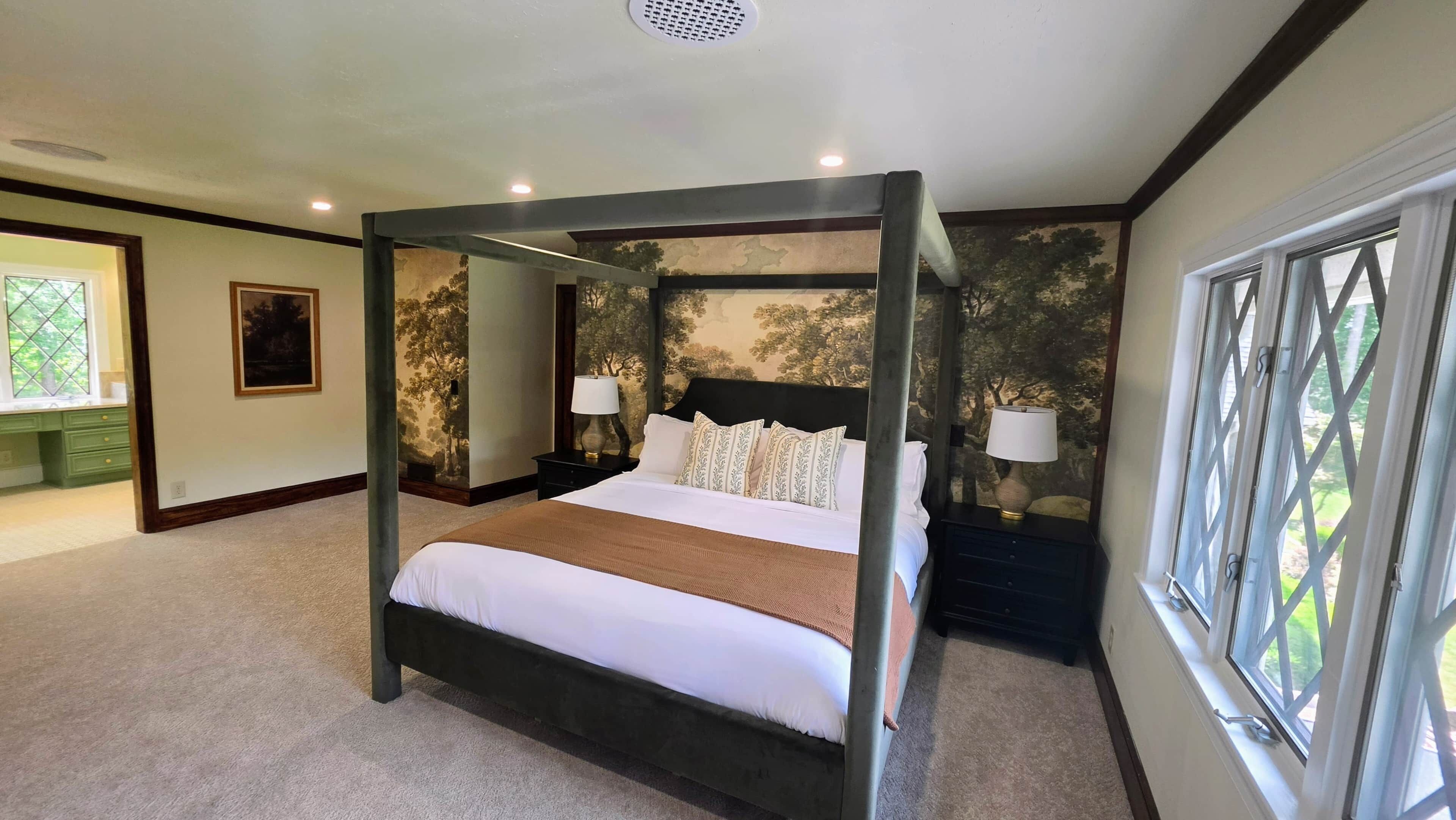 A four-poster bed with a white bedspread and multiple pillows, flanked by nightstands with lamps, in a room with a large window and a mural on the wall.