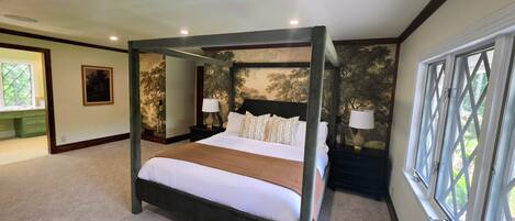 A four-poster bed with a white bedspread and multiple pillows, flanked by nightstands with lamps, in a room with a large window and a mural on the wall.