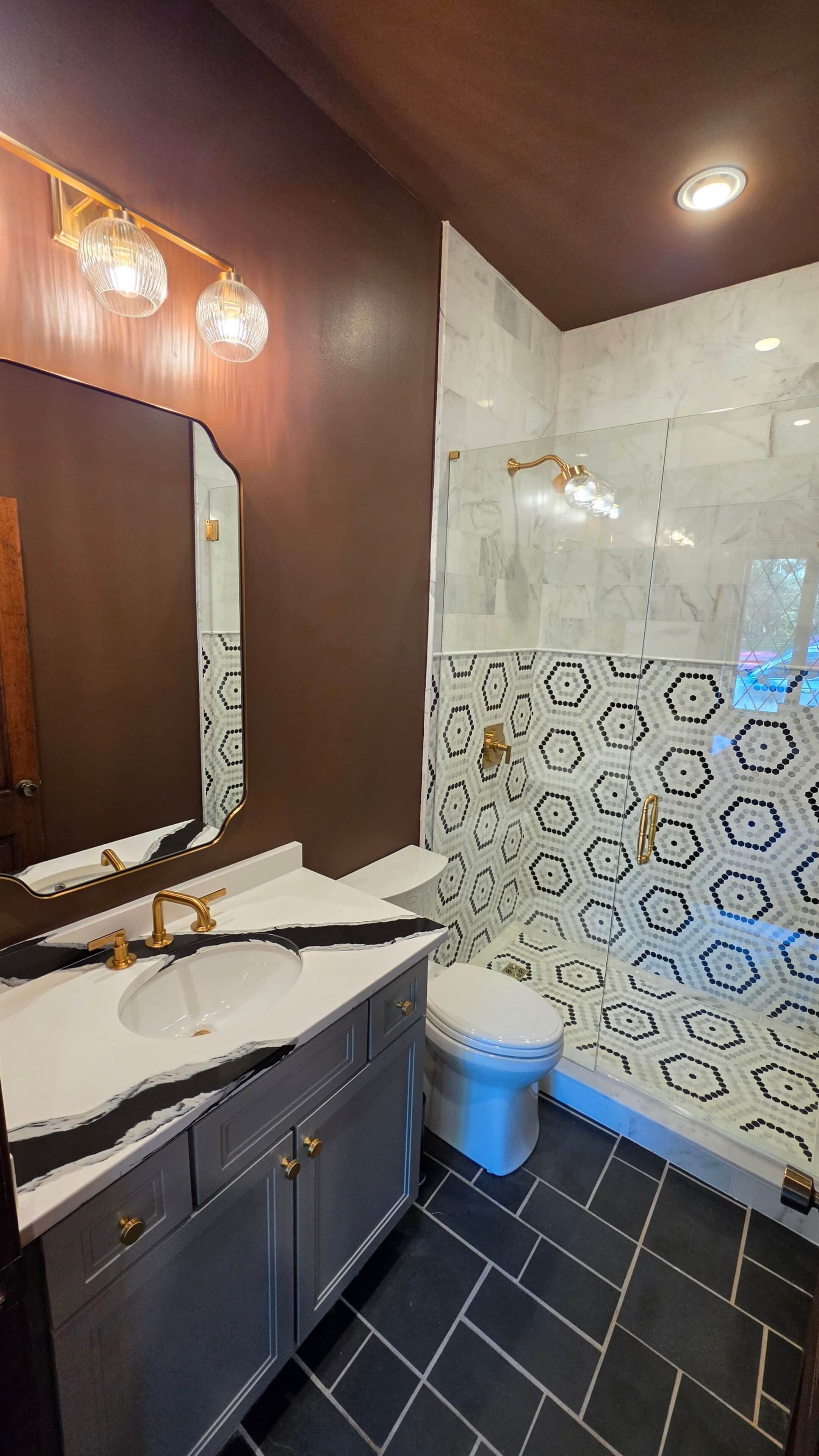 Une salle de bain dotée d’un grand miroir, d’un lavabo avec des robinets dorés, de toilettes et d’une douche dont le mur est recouvert de carreaux à motifs.