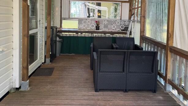 A covered outdoor area with a wooden deck, wicker furniture, and a view of a house through a window.