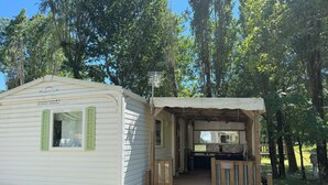 A small, single-story mobile home with a porch and a small attached structure, surrounded by trees.