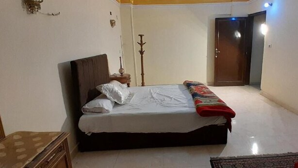 A bedroom with a bed, a bedside table, a lamp, a wardrobe, and a red chair. - Happy hotel pyramids inn  (Giza Governorate)