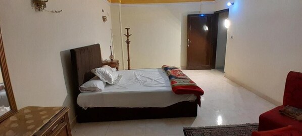 A bedroom with a bed, a bedside table, a lamp, a wardrobe, and a red chair. - Happy hotel pyramids inn  (Giza Governorate)