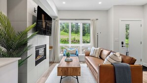 A modern living room with a fireplace, a sofa, a coffee table, and a view of a garden through large windows.