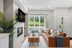 Un salon moderne avec une cheminée, un canapé, une table basse et une vue sur un jardin grâce  à de grandes fenêtres.