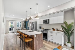 Une cuisine moderne dotée d’un îlot central, d’appareils électroménagers en acier inoxydable, de luminaires suspendus et offrant une vue sur l’extérieur.