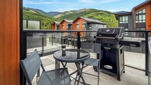 A balcony with a glass railing, a round table, two chairs, and a barbecue grill.
