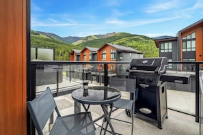 A balcony with a glass railing, a round table, two chairs, and a barbecue grill. - Ridgeline | Slope-Side | Hot Tub | Resort Access (Panorama)