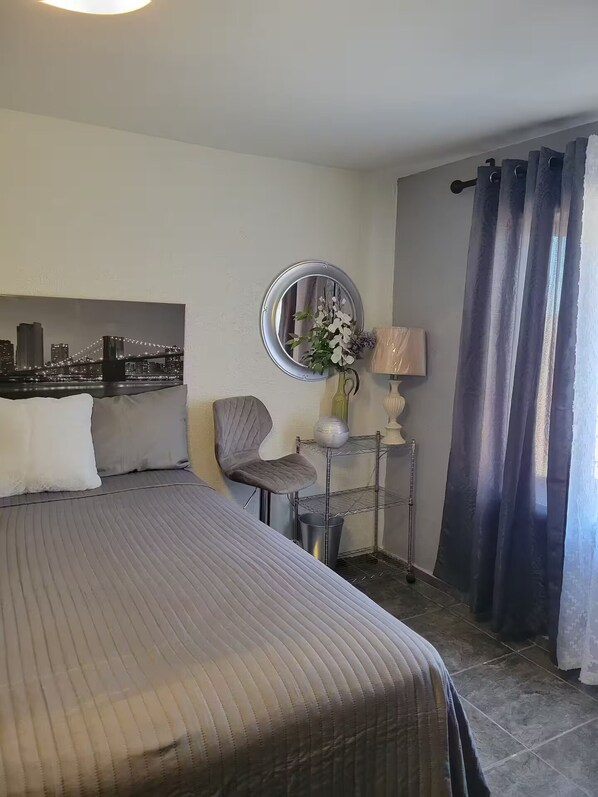 A hotel room with a bed, a chair, a small table with a lamp, a mirror, and a cityscape view.