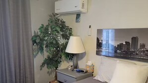 Una camera d'albergo con un letto, un comodino con una lampada, una pianta in vaso, un condizionatore d'aria e un quadro raffigurante un panorama cittadino.