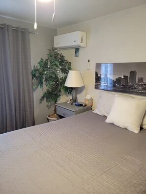 Hotellihuone, jossa on sänky, yöpöytä lampun kanssa, ruukkukasvi, ilmastointilaite ja kaupunkimaisemaa esittävä taulu seinällä.