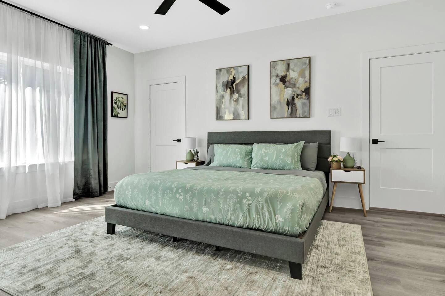 A modern bedroom with a bed, two nightstands, a large window with curtains, and abstract wall art.