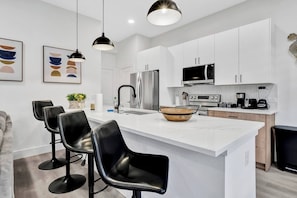 A modern kitchen with white cabinetry, a central island with stools, a stainless steel refrigerator, a microwave, and a coffee station. - Dallas Escape w/ Rooftop + Yard • Sleeps 10 (Dallas)