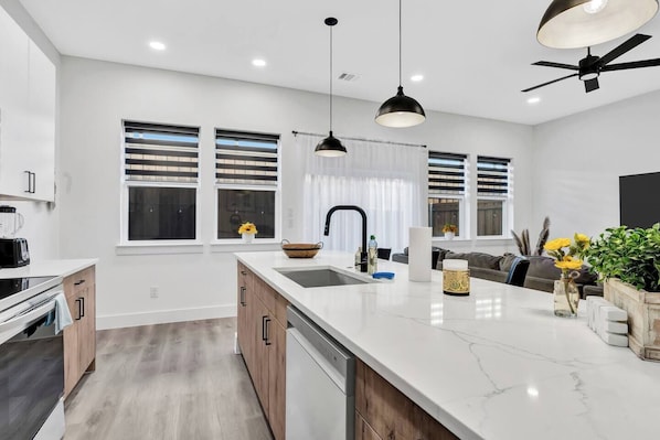 A modern kitchen with a marble countertop, wooden cabinets, a built-in dishwasher, and a large window with blinds. - Dallas Escape w/ Rooftop + Yard • Sleeps 10 (Dallas)
