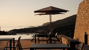 An outdoor seating area with tables and chairs, a stone wall, and a view of the water and hills. - KHAI SYVOTA (Igoumenitsa)